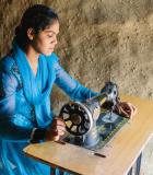 A woman in a blue dress works on a garment at a sewing machine. 