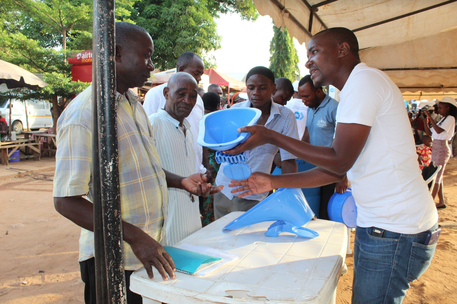 A man in a white t-shirt and jeans standing behind a table demonstrates a blue plastic latrine pan to three other men