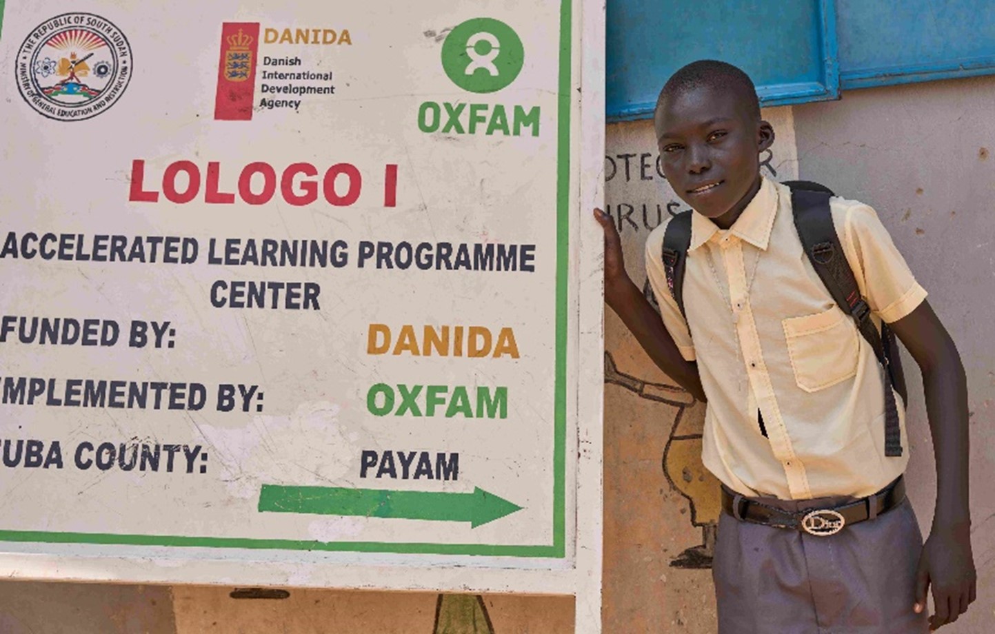 A young boy stands in front of his school's information sign. He is wearing a pale yellow button up shirt, dark blue pants, and a backpack.