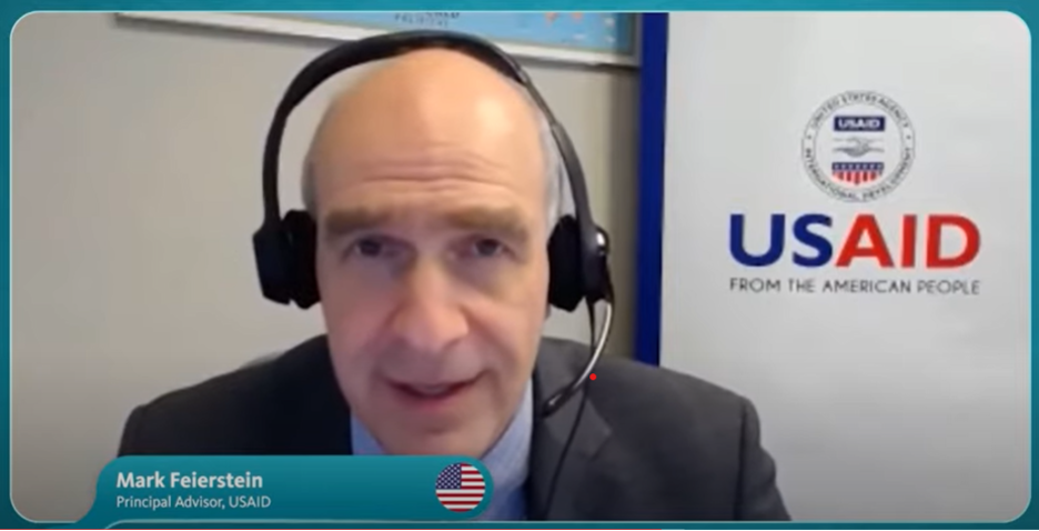 "a screenshot of Mark Feierstein wearing a headset with the USAID logo in the background"