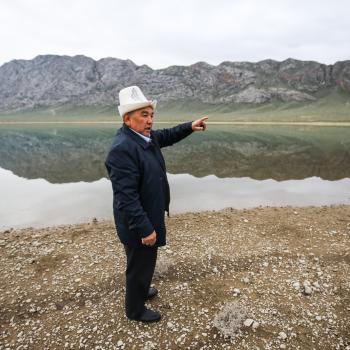 A man in a white hat and dark coat points into the distance standing on the banks of a body of water. There are mountains in the background.