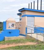 A cement building with steps and an access ramp, painted in bright blue and tan colors.  A small round tower has a mural reminding children to wash their hands after using the toilet.
