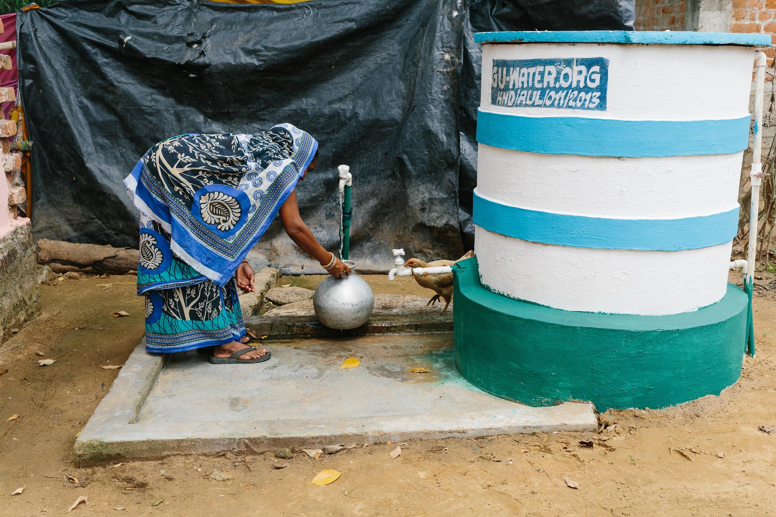 A woman in a patterned blue and white sari bends down to collect water from a pump in the ground.
